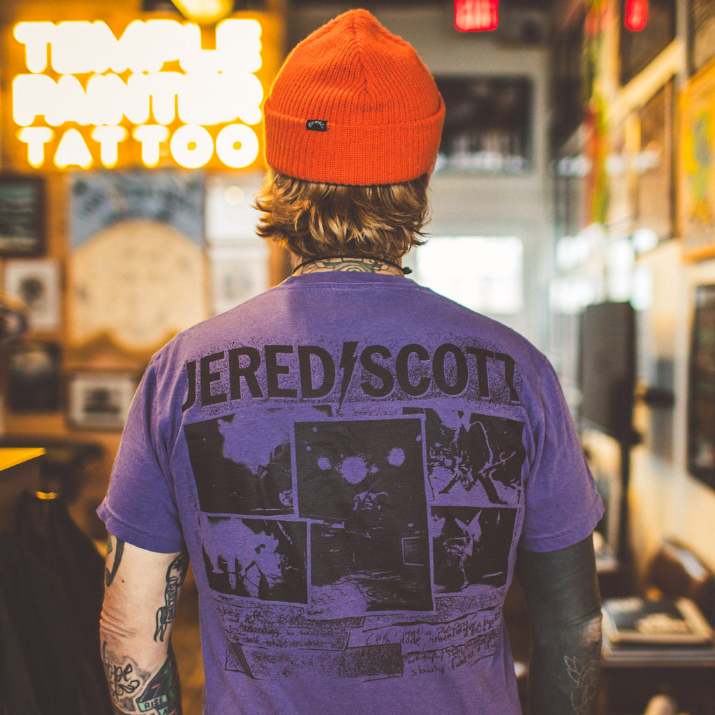 Person wearing a purple t-shirt with 'Jered/Scott' design in a tattoo parlor.
